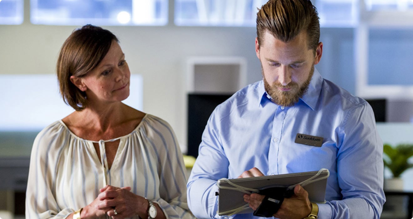 Two people in an office environment are standing side-by-side, looking at a mobile tablet together.