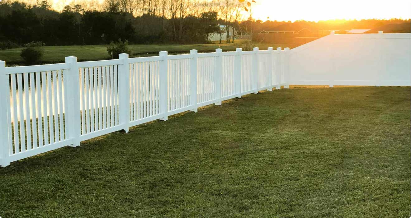 A scenice picture of a yard wrapped in a white vinyl fence. Behind the fence is a small pond and a setting sun.